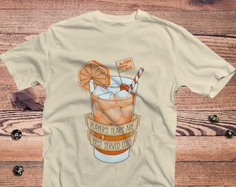 Puede incluir: Una camiseta beige con un gráfico de un vaso de zumo de naranja con cubitos de hielo y una rodaja de naranja. El texto "Player's Tears Are Best Served Cold" está impreso en una pancarta alrededor del vaso. Una pequeña bandera con el texto "The DM Always Wins" está clavada en la rodaja de naranja.