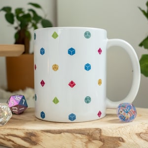 Può includere: Tazza in ceramica bianca con un motivo ripetitivo di dadi colorati. I dadi sono di varie forme e colori, tra cui rosso, blu, verde, giallo e viola.