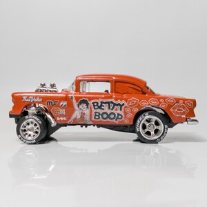May include: A red toy car with a Betty Boop design. The car has a black and white design with the words 'Betty Boop' and 'True Value' on the side. The car has silver wheels with black tires.