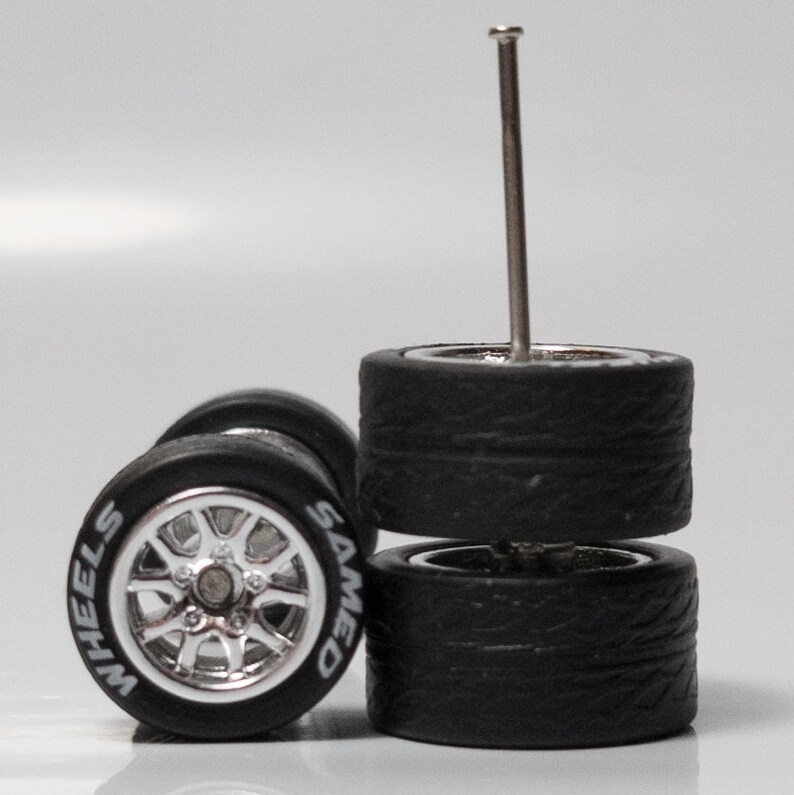 May include: Two sets of black rubber tires with silver rims. The tires have a tread pattern and the rims have the words 'WHEELS' and 'SAMED' printed on them. A silver metal pin is sticking out of the top of the tires.