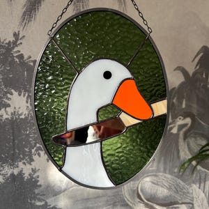 Puede incluir: Una ventana de vitral con una oca blanca con un pico naranja que sostiene un cuchillo en su pico. La oca est&aacute; sobre un fondo verde.
