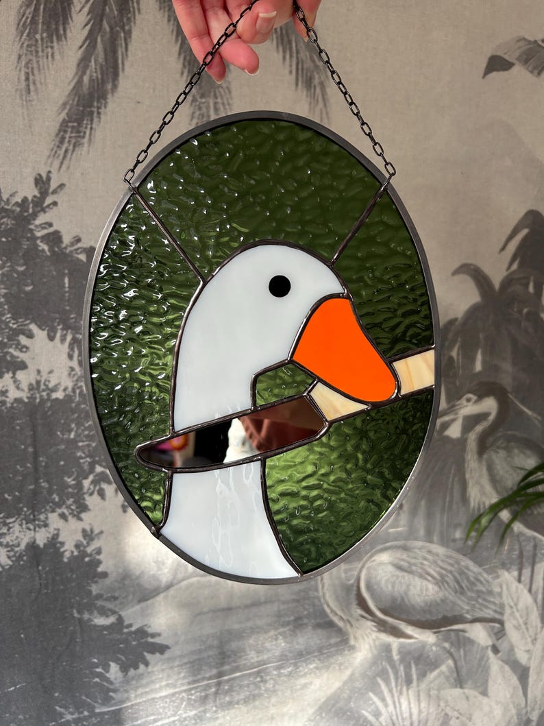 Puede incluir: Una ventana de vitral con una oca blanca con un pico naranja que sostiene un cuchillo en su pico. La oca est&aacute; sobre un fondo verde.