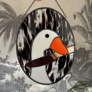 Puede incluir: Una ventana de vitral colgada de una cadena, que representa un ganso blanco con un pico naranja y un ala marr&oacute;n. El ganso est&aacute; colocado sobre un fondo marmoleado en blanco y negro.