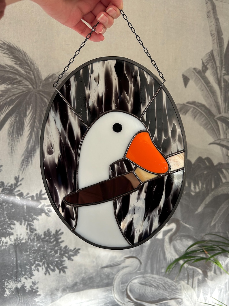 Puede incluir: Una ventana de vitral colgada de una cadena, que representa un ganso blanco con un pico naranja y un ala marr&oacute;n. El ganso est&aacute; colocado sobre un fondo marmoleado en blanco y negro.