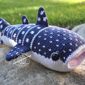 May include: A crocheted whale shark plush toy with a blue and white spotted pattern. The plush toy has a pink mouth and black eyes. The toy is sitting on a rock.