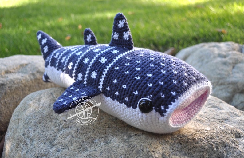 May include: A crocheted whale shark plush toy with a blue and white spotted pattern. The plush toy has a pink mouth and black eyes. The toy is sitting on a rock.