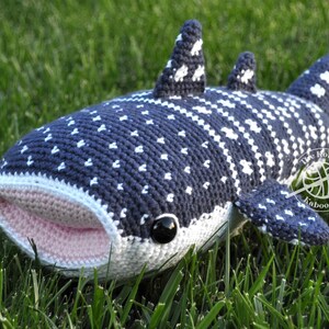 May include: A crocheted whale shark plush toy with a pink mouth, black eyes, and a white and blue spotted pattern. The toy is lying on green grass.