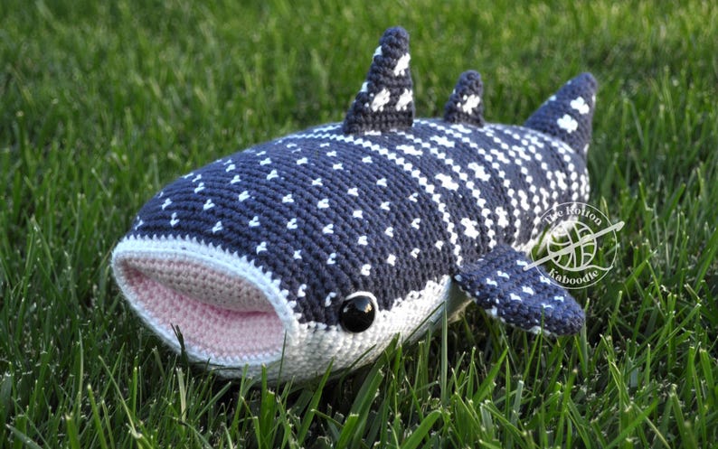 May include: A crocheted whale shark plush toy with a pink mouth, black eyes, and a white and blue spotted pattern. The toy is lying on green grass.