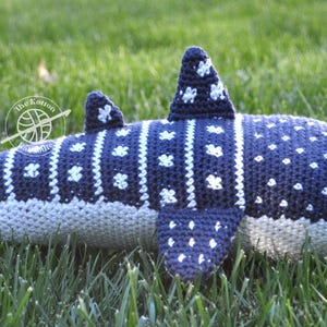 May include: A crocheted whale shark plush toy with a blue and white spotted pattern. The plush toy is lying on a bed of green grass.