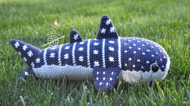 May include: A crocheted whale shark plush toy with a blue and white spotted pattern. The plush toy is lying on a bed of green grass.
