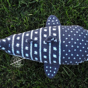 May include: A crocheted whale shark plush toy with a gray and white pattern. The plush toy is lying on a bed of green grass.