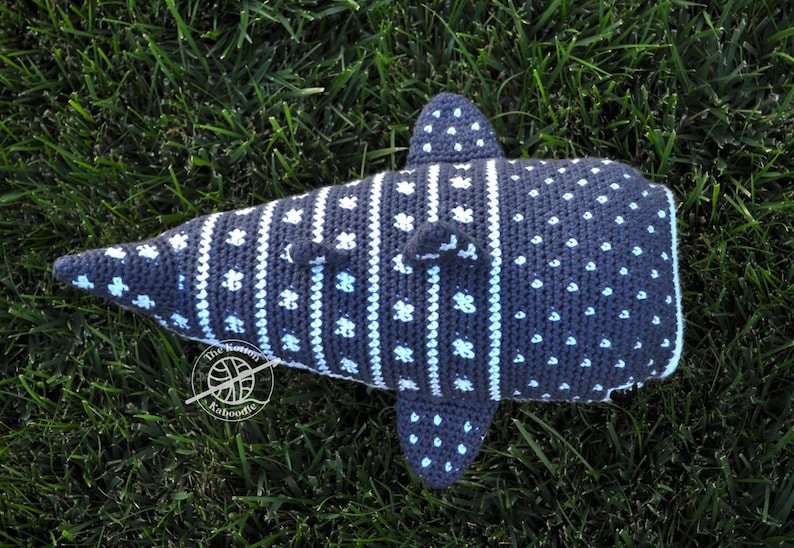 May include: A crocheted whale shark plush toy with a gray and white pattern. The plush toy is lying on a bed of green grass.