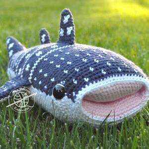 May include: A crocheted whale shark plush toy with a light blue and white patterned body, pink mouth, and black eyes. The plush toy is lying on green grass.