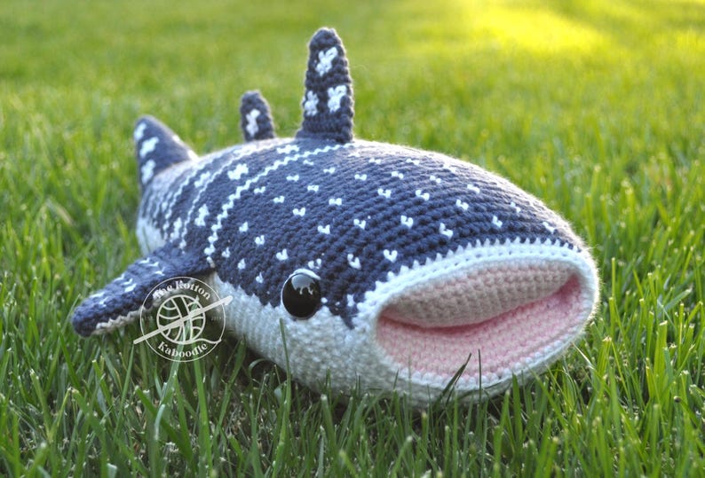 May include: A crocheted whale shark plush toy with a light blue and white patterned body, pink mouth, and black eyes. The plush toy is lying on green grass.