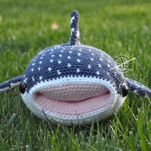 May include: A crocheted whale shark plush toy with a pink mouth and white spots on a blue body. The toy is lying on green grass.