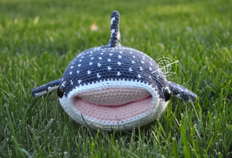 May include: A crocheted whale shark plush toy with a pink mouth and white spots on a blue body. The toy is lying on green grass.