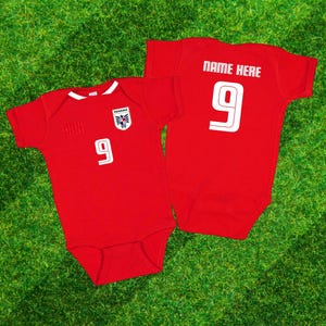 May include: Two red baby bodysuits with white accents, featuring the number 9. One has a crest and the word "PANAMA". The other has "NAME HERE" above the number 9. The bodysuits are on a green grass background.