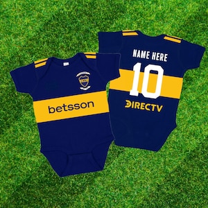 May include: Two navy blue baby onesies with yellow stripes and accents. One has the text "betsson" and a crest, the other has "NAME HERE", the number "10", and "DIRECTV". The onesies are on a green grass background.