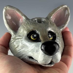 May include: A ceramic wolf head sculpture with gray fur, pink-lined ears, and a black nose. The wolf has yellow eyes and is held in a hand. The sculpture is a decorative item.
