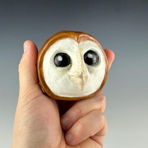 May include: A handcrafted owl figurine with a round shape. The owl has large, black eyes, a white face, and a brown head. The figurine is held in a hand, showcasing its size and detail. The owl's beak is a light beige color.