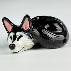 May include: A ceramic figurine of a sleeping husky dog. The dog is black and white with pink inner ears and a black nose. The figurine is curled up, with its eyes closed, and appears to be resting.