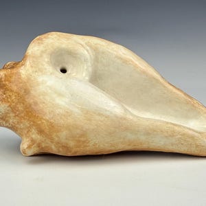 May include: A ceramic sculpture in the shape of a seashell, with a light beige and brown color palette. The sculpture has a smooth, curved design with a small hole at the top and a rounded end.