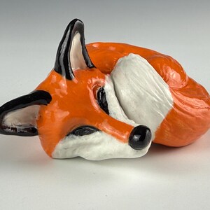 May include: A ceramic fox figurine in a curled-up sleeping position. The fox has orange fur with white accents on its chest and tail. The ears and nose are black. The figurine is on a white surface.