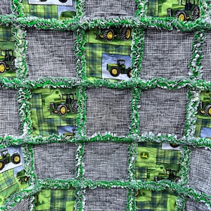 May include: A green and gray patchwork quilt with a John Deere tractor theme. The quilt is made of squares with different John Deere tractor designs, including a tractor with a plow, a tractor with a trailer, and a tractor with a logo. The quilt is edged with green and white fringe.