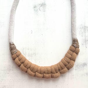 May include: A beige and tan knitted necklace with a silver clasp. The necklace features a thick, textured design with a rounded shape. The upper portion is a light beige color, transitioning to a darker tan at the bottom.