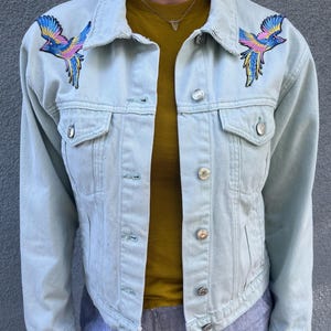 Könnte beinhalten: Hellblaue Jeansjacke mit gestickten Vogelmotiven in Blau, Gelb und Rosa. Die Jacke hat silberne Knöpfe und zwei Vordertaschen. Darunter ist ein gelbes T-Shirt zu sehen.