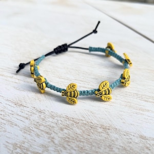 May include: Adjustable bracelet featuring yellow bee charms on a teal cord. The bracelet has a black sliding knot closure. The bees have black stripes and leaf details. The bracelet is displayed on a white wooden surface.