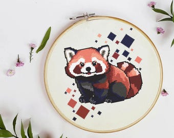 May include: A cross-stitch embroidery hoop featuring a red panda design. The panda is rendered in shades of red, black, and white, with a pixelated style. Geometric shapes in red and blue surround the panda on a white background.