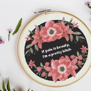 May include: A cross-stitch embroidery hoop with a black background and the text "If pain is beauty, I'm a pretty bitch." The design features pink flowers and green leaves. The wooden hoop is round.