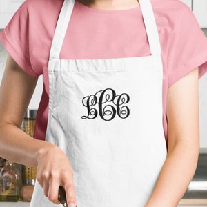 May include: A white apron with a monogrammed design in black. The apron has a pocket on the front. A person is wearing the apron and is chopping vegetables on a wooden cutting board.