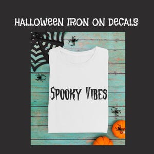 May include: White t-shirt with the text "Spooky Vibes" in black, displayed on a teal wooden surface with Halloween decorations. The decorations include a black spider web, plastic spiders, and small orange pumpkins.