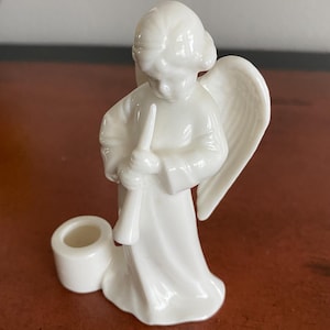 May include: A white ceramic angel figurine with a trumpet and a candle holder. The angel has detailed wings and is wearing a flowing robe. The figurine is on a brown surface.