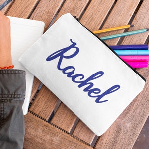 May include: White canvas pencil case with a blue cursive name "Rachel" printed on it. The case is open and has several colored pencils and pens inside.