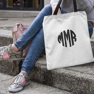 May include: A white canvas tote bag with black handles and a black monogram of the letters "MMR" in a circle.