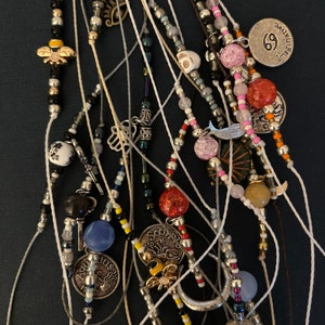 May include: A collection of beaded bracelets with various charms, including zodiac signs, a bee, a crescent moon, and a skull. The bracelets are made with white string and feature colorful beads.