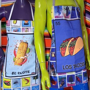 May include: Two aprons featuring colorful Loteria card graphics. The left apron displays a large corn cob with the text EL ELOTE. The right apron features a large taco graphic with the text LOS TACOS and the number 55. Both have white neck straps.