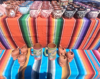 May include: A selection of handcrafted ceramic mugs and skull-shaped planters displayed on a vibrant striped textile. The mugs showcase floral and geometric patterns in brown, blue, and white. The planters are a terracotta colour.