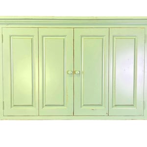 May include: A light green wall-mounted cabinet with four doors. The cabinet has a rectangular shape with a decorative top molding and small round knobs. The doors have recessed panels and a distressed finish.