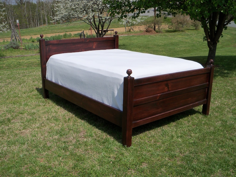 May include: A dark brown wooden bed frame with a high headboard and a lower footboard, both featuring square posts topped with spherical finials. The bed is dressed with a plain white sheet covering the mattress surface, showcasing a simple design.