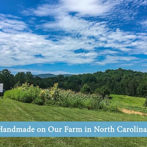 Landscape photo of March Legend’s North Carolina farm featuring rolling green fields, sunflowers, trees, and mountains under a bright blue sky with the text “Handmade on Our Farm in North Carolina.