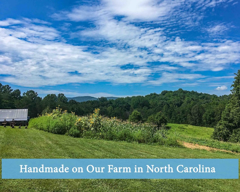 Landscape photo of March Legend’s North Carolina farm featuring rolling green fields, sunflowers, trees, and mountains under a bright blue sky with the text “Handmade on Our Farm in North Carolina.