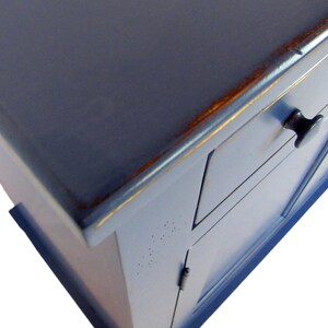 K&ouml;nnte beinhalten: Ein blauer, lackierter Holzschrank mit zwei T&uuml;ren und einer Schublade. Der Schrank hat eine abgenutzte Oberfl&auml;che und schwarze Metallkn&ouml;pfe.