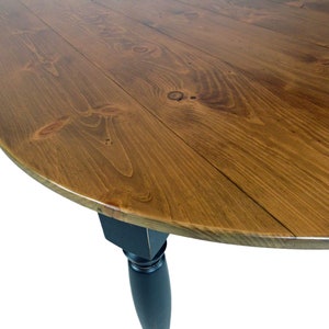 May include: A round dining table featuring a natural brown wood grain tabletop with visible knots and a smooth finish. The table is supported by a single, solid black turned leg that tapers toward the bottom, providing a stable base for the surface.