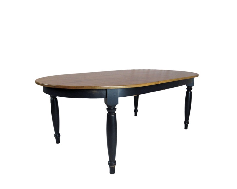 May include: An oval dining table featuring a smooth, medium-brown wood grain tabletop and a black-painted apron. The table is supported by four turned, tapered legs finished in black, showcasing a classic silhouette with a two-tone color scheme.
