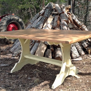 May include: A rectangular wooden dining table with a natural light brown wood grain top and a cream-colored base. The table features a trestle-style support structure with a horizontal stretcher bar connecting two curved, wide-footed vertical legs.