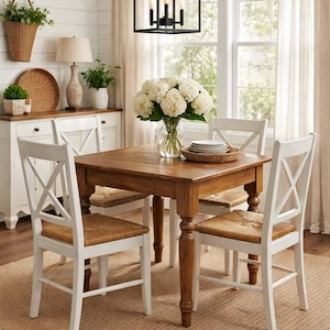 May include: A dining room set featuring a square wooden table with four white chairs. The chairs have woven seats and an X-shaped back. A vase of white flowers sits on the table. The room has a neutral color palette.